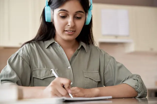 indian-girl-wearing-headphones-writing-homework-listening-audio-course-home_203461-1202.webp