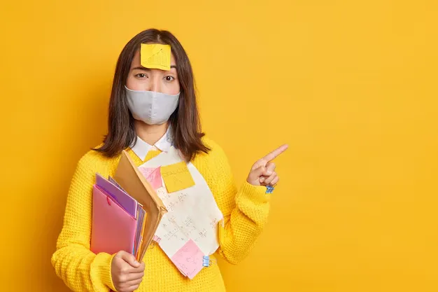 serious-brunette-asian-woman-has-sticker-head-tries-learn-information-pass-exam-successfully-wears-disposable-mask-prevent-coronavirus-points-blank-empty-space_273609-46394.webp
